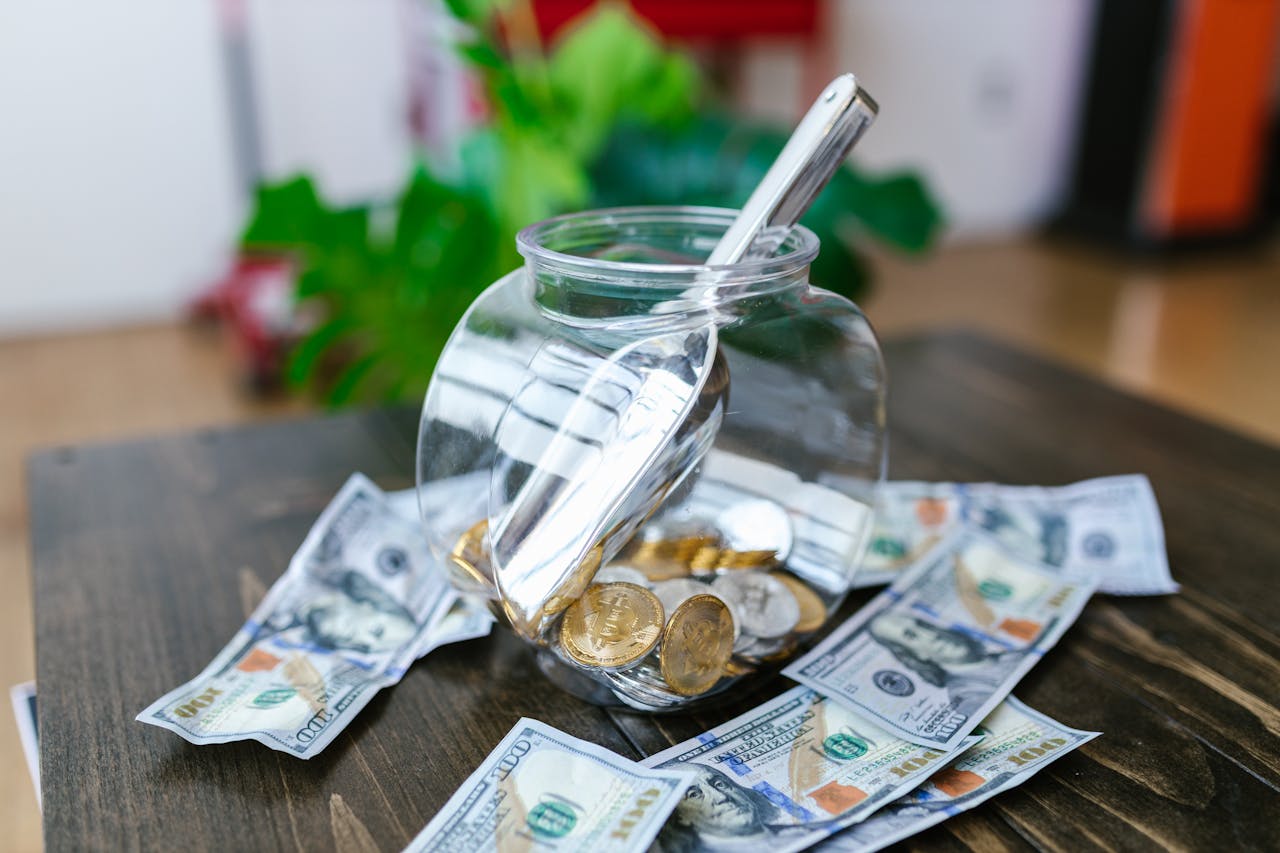 A transparent jar filled with coins and surrounded by dollar bills on a wooden table.